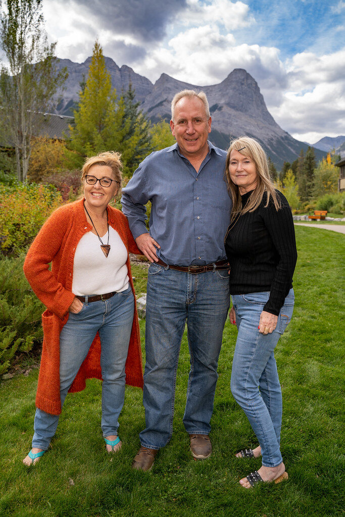 A group shot of Teresa Mullen, Ross Jansen and Ann Ricord of Spring Creek Real Estate in Canmore, standing in front of green foliage with mountains in the background.
